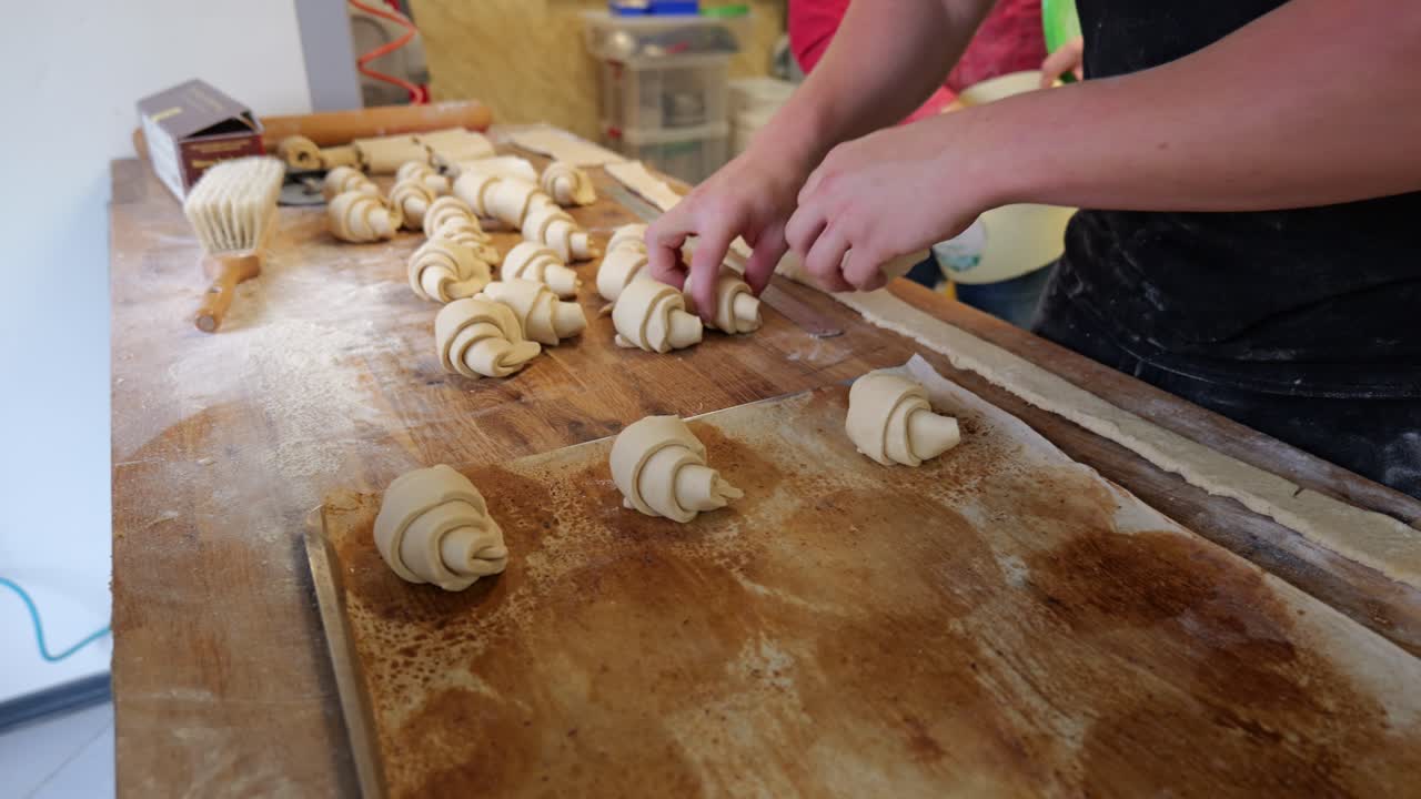 Zbliżenie na dłonie piekarza układającego surowe croissanty na metalową blachę do pieczenia.