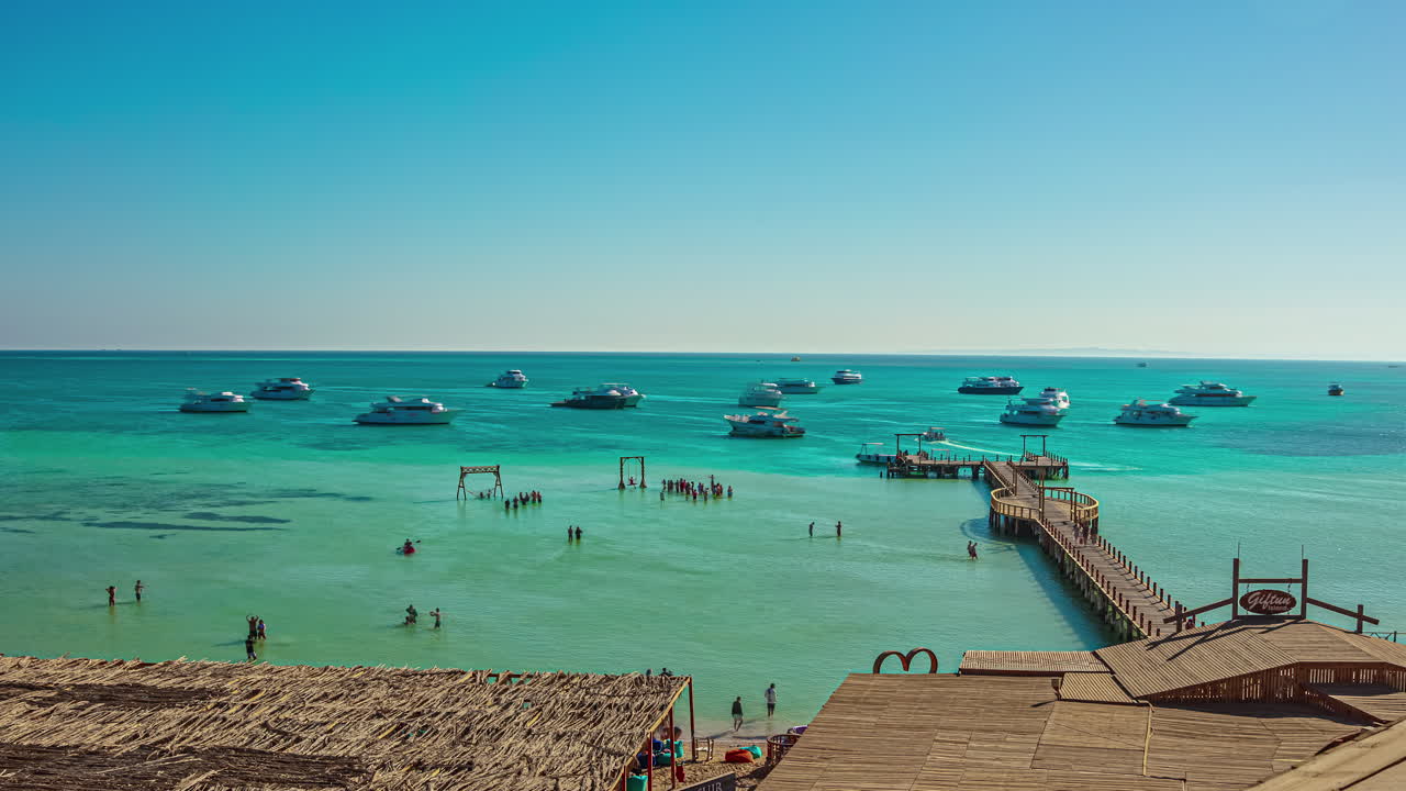 Timelapse shot of busy beachside with people bathing and yachts docked on a sunny day