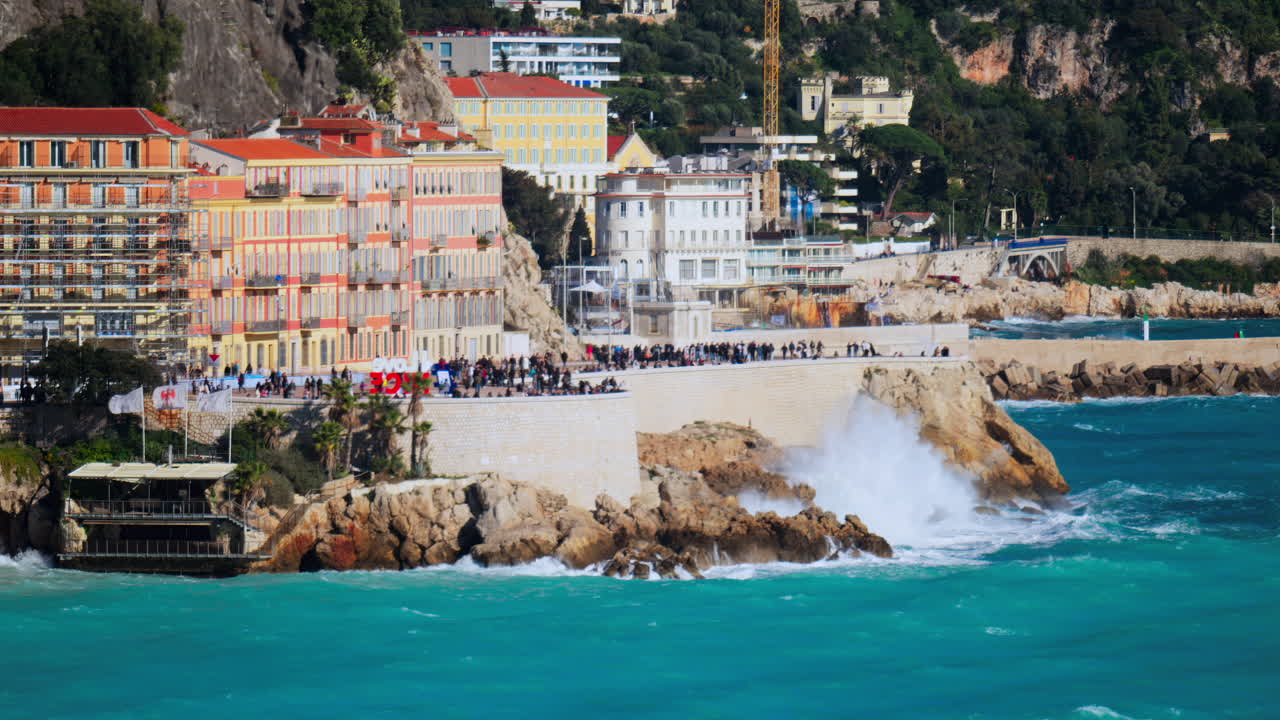 Nice, France - March 17, 2025: View of people walking on the coast of the city with water crashing on the rocks and the buildings on the background