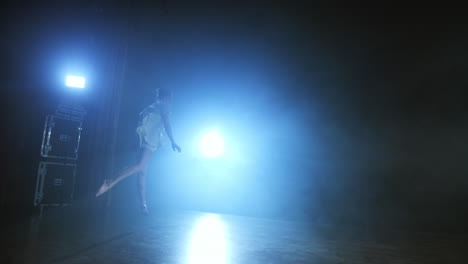 a young girl in a white dress dances a modern ballet spins on the leg and jumps on the stage with smoke against the blue spotlights.