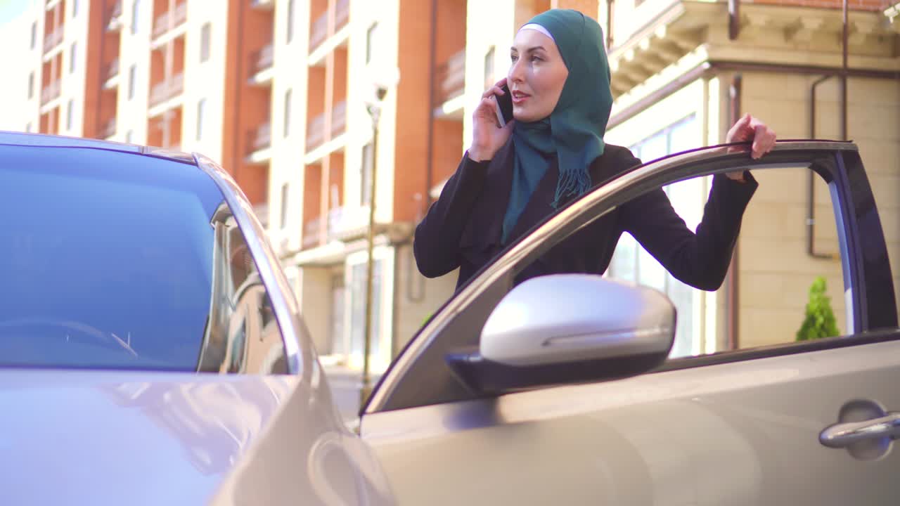 joven musulmana en hijab hablando por teléfono y sentada en el coche en casa