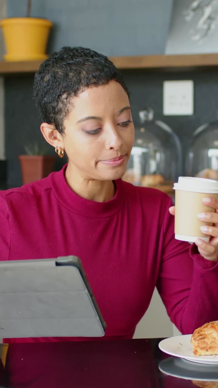 Video verticale: Una donna in bordeaux che tocca un tablet grigio per leggere e afferra una tazza in un caffè, con un croissant.