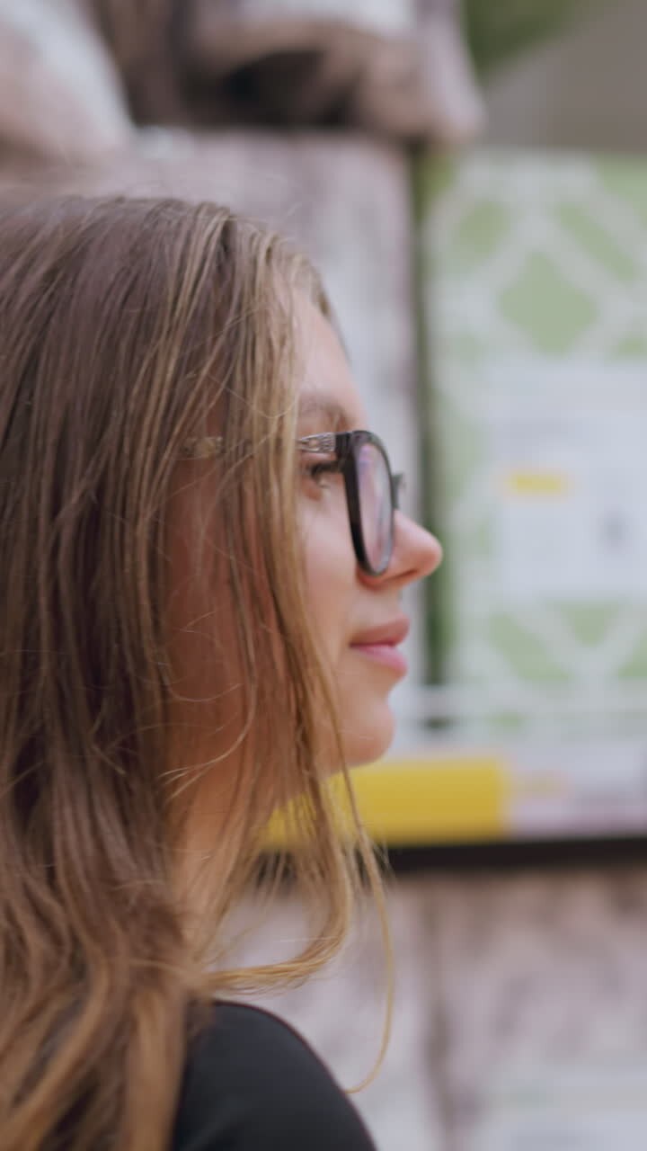 Head view of woman with long brown hair wearing black top and glasses browsing supermarket aisle looking at household products on shelves, background blurred with various packaged goods and labels