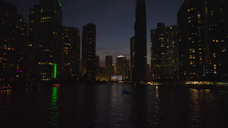 Low-angle-view-of-tall-buildings-with-lighted-windows-around-river.-Forwards-fly-above-water-surface-at-night.-Miami,-USA