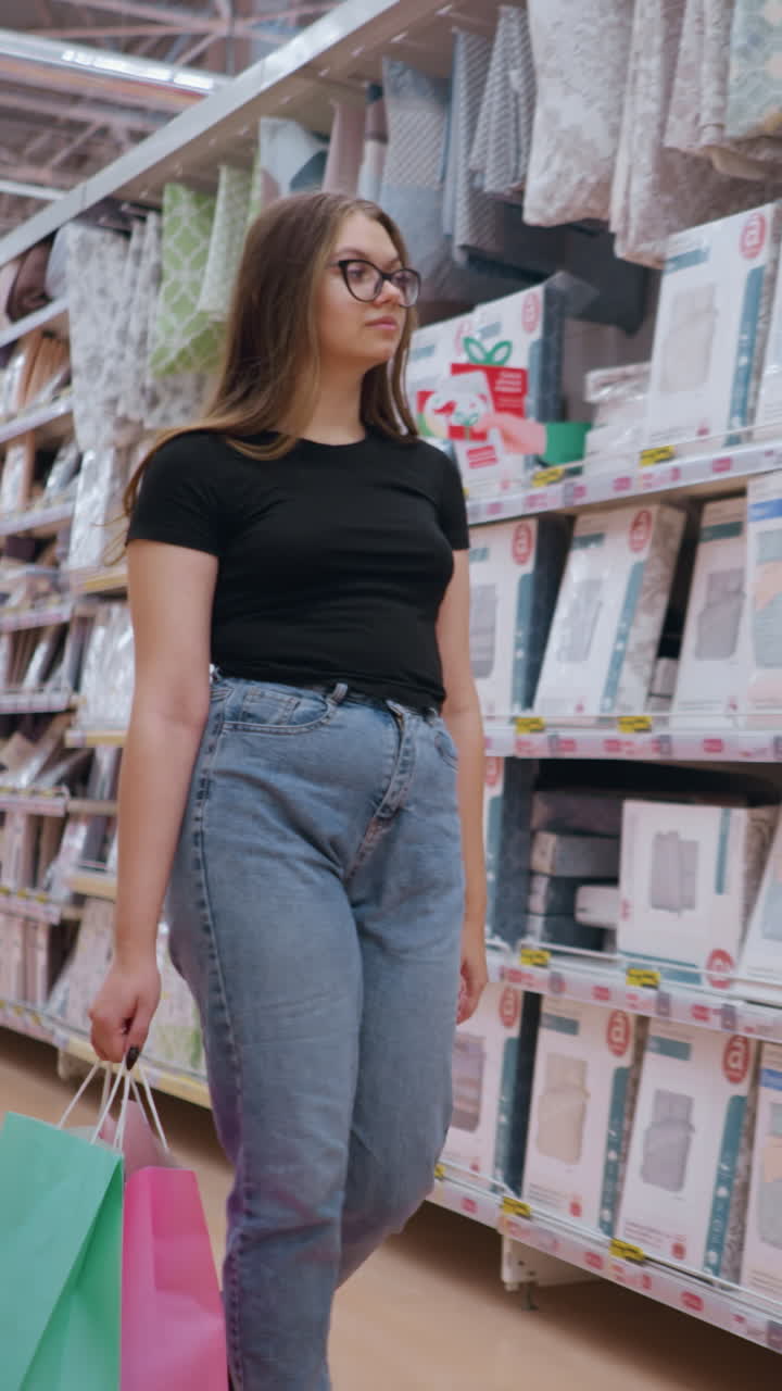 mujer joven con gafas, camiseta negra y vaqueros camina por las estanterías de las tiendas llenas de ropa de cama, sosteniendo bolsas de compras coloridas mientras mira a su alrededor, entorno de venta al por menor con otros compradores