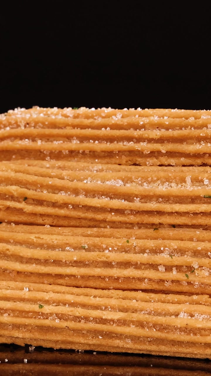 Churros dorati impilati a piramide ruotano e si ingrandiscono sotto un'illuminazione da studio drammatica, evidenziando i cristalli di zucchero e la consistenza croccante su uno sfondo nero.