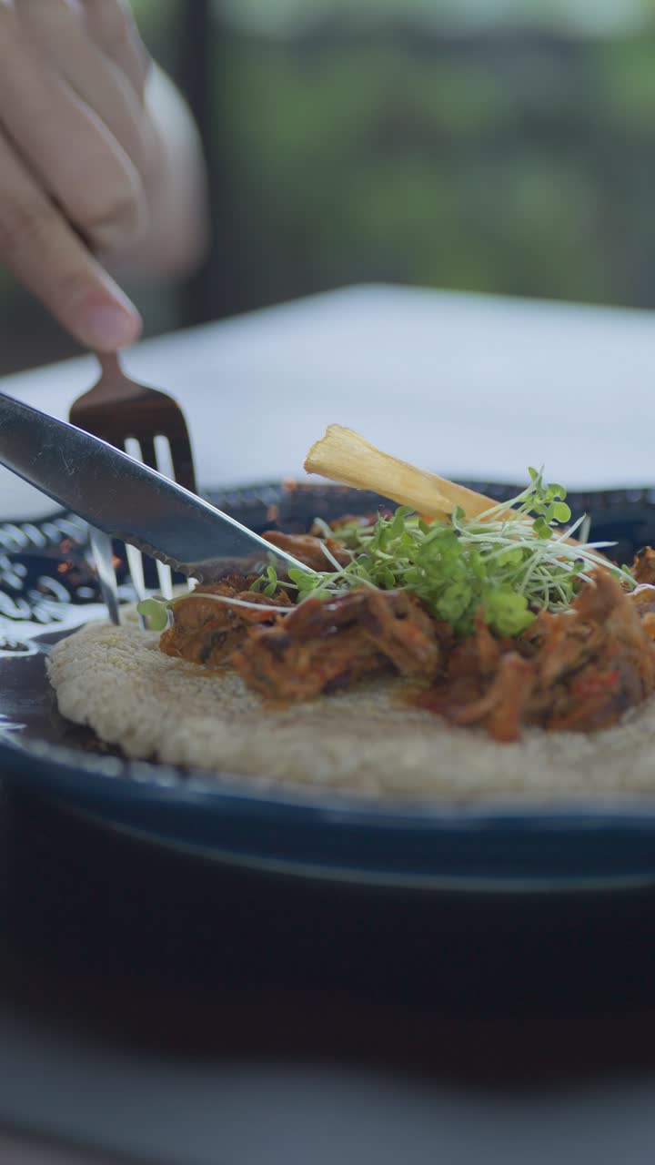 Una persona sta mangiando con una forchetta e un telefono cellulare in mano. Il cibo è un mix di carne e verdure, con alcune foglie verdi sopra. La scena è casuale e rilassata.