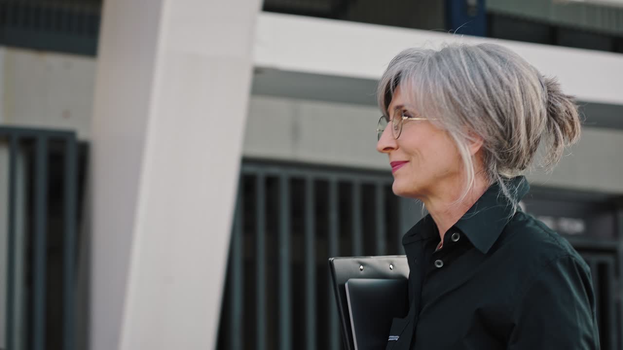 Beautiful gray-haired woman manager walks near the office center smiling, looks at her wrist watch