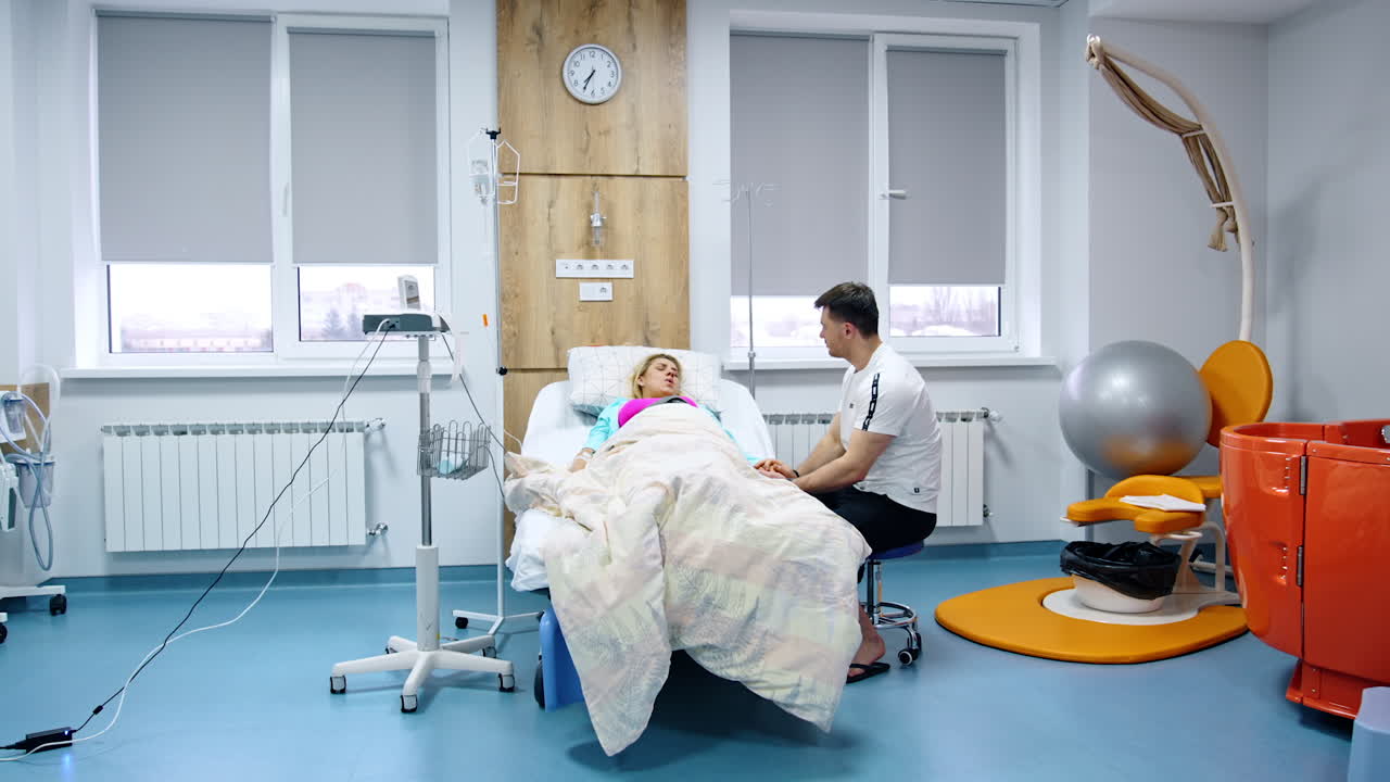 Loving husband sits at the bed of his pregnant wife. Blonde woman suffering from painful contractions. Modern hospital ward backdrop.