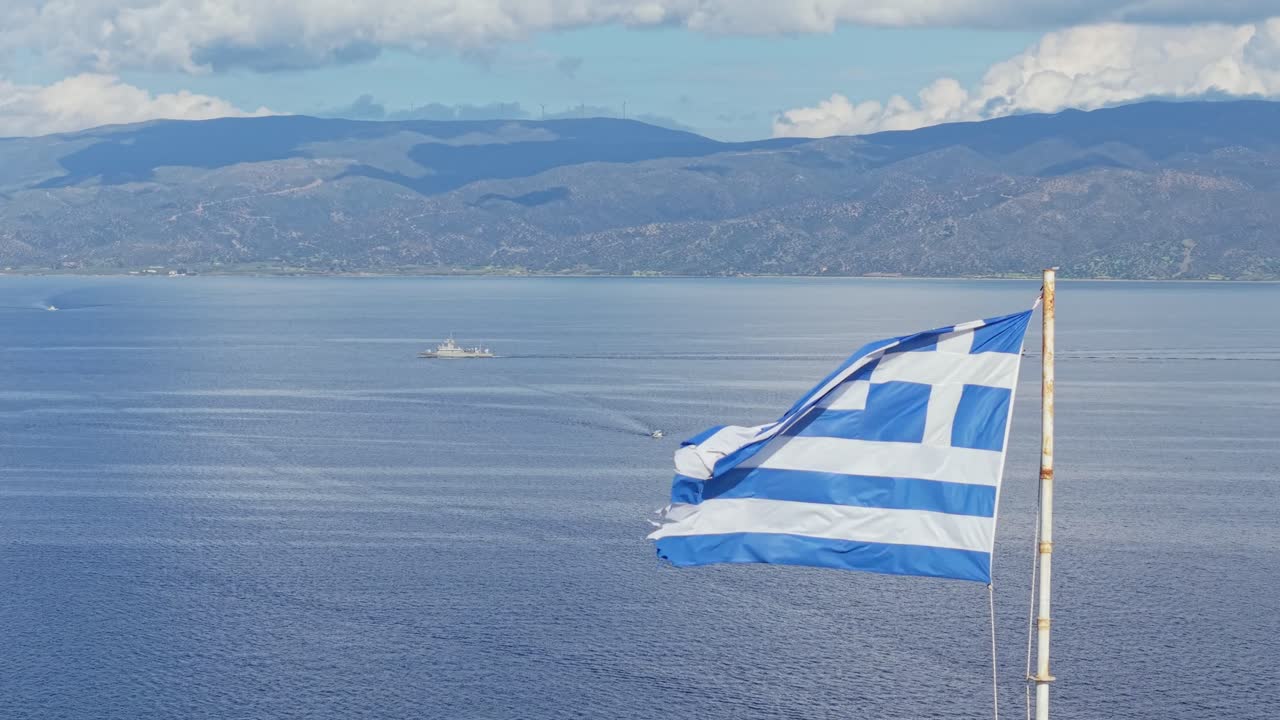 En drone kredser om det græske flag med det dybblå Ægæiske Hav i baggrunden, mens et græsk flådeskib passerer forbi og symboliserer national stolthed og maritim arv.