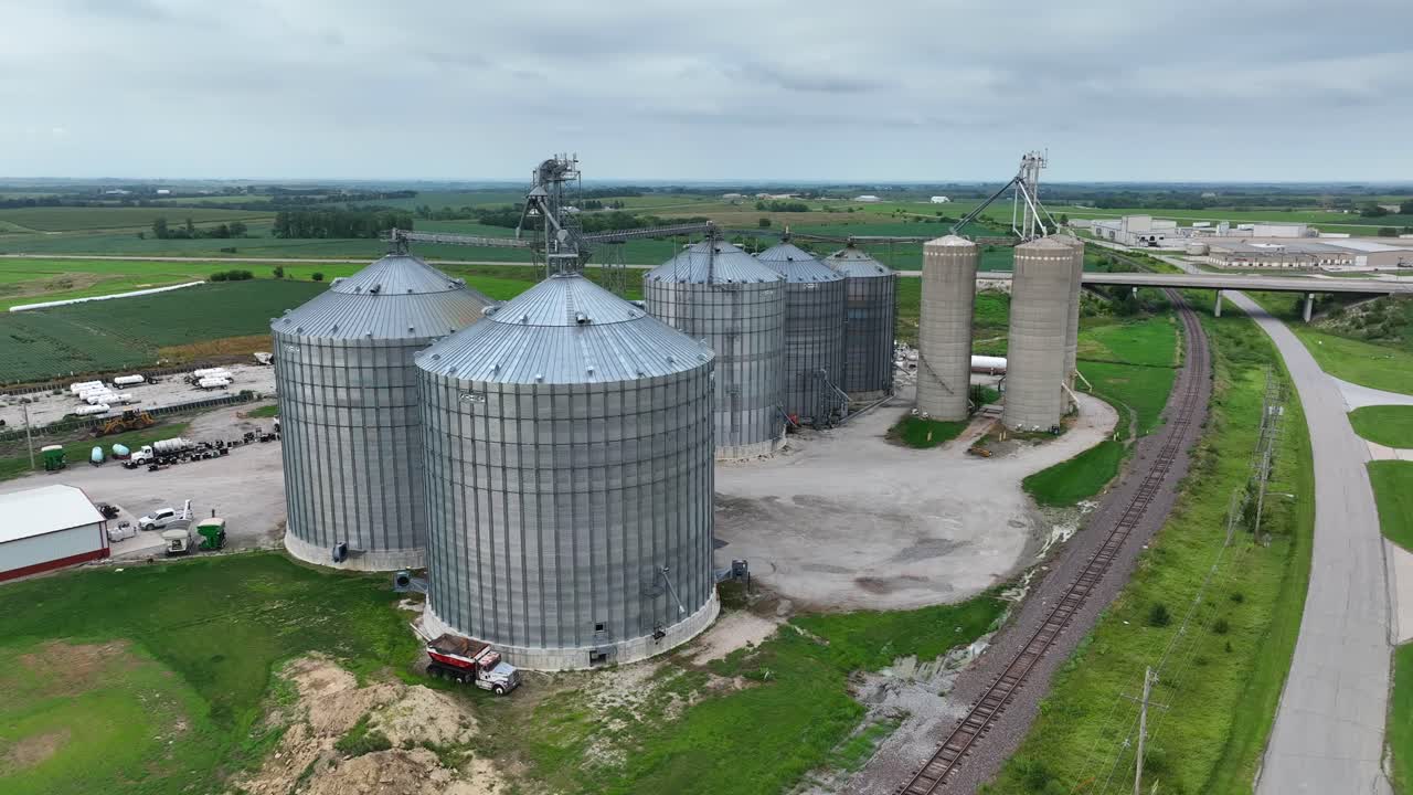 silos de grano en el medio oeste, con vastas tierras de cultivo, una vía férrea, y una carretera cerca bajo un cielo nublado
