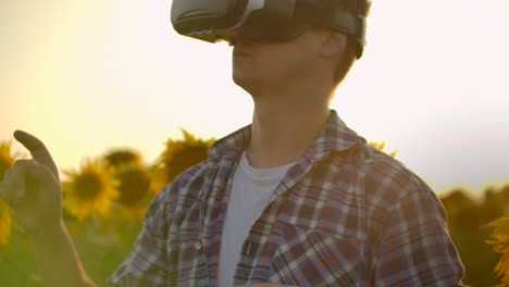 a male in plaid shirt and jeans uses vr glasses on the field with sunflowers for scientific article. these are new technologies at sunset.