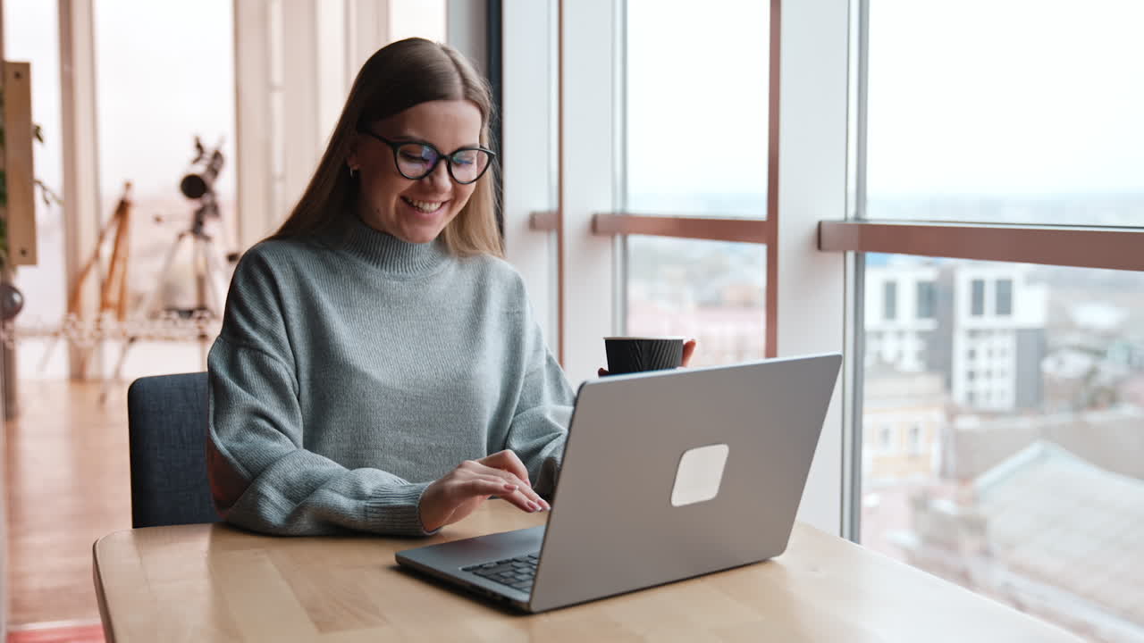Cheerful lady in glasses having conversation via internet. Woman with coffee cup finishes conversation and looks at window.