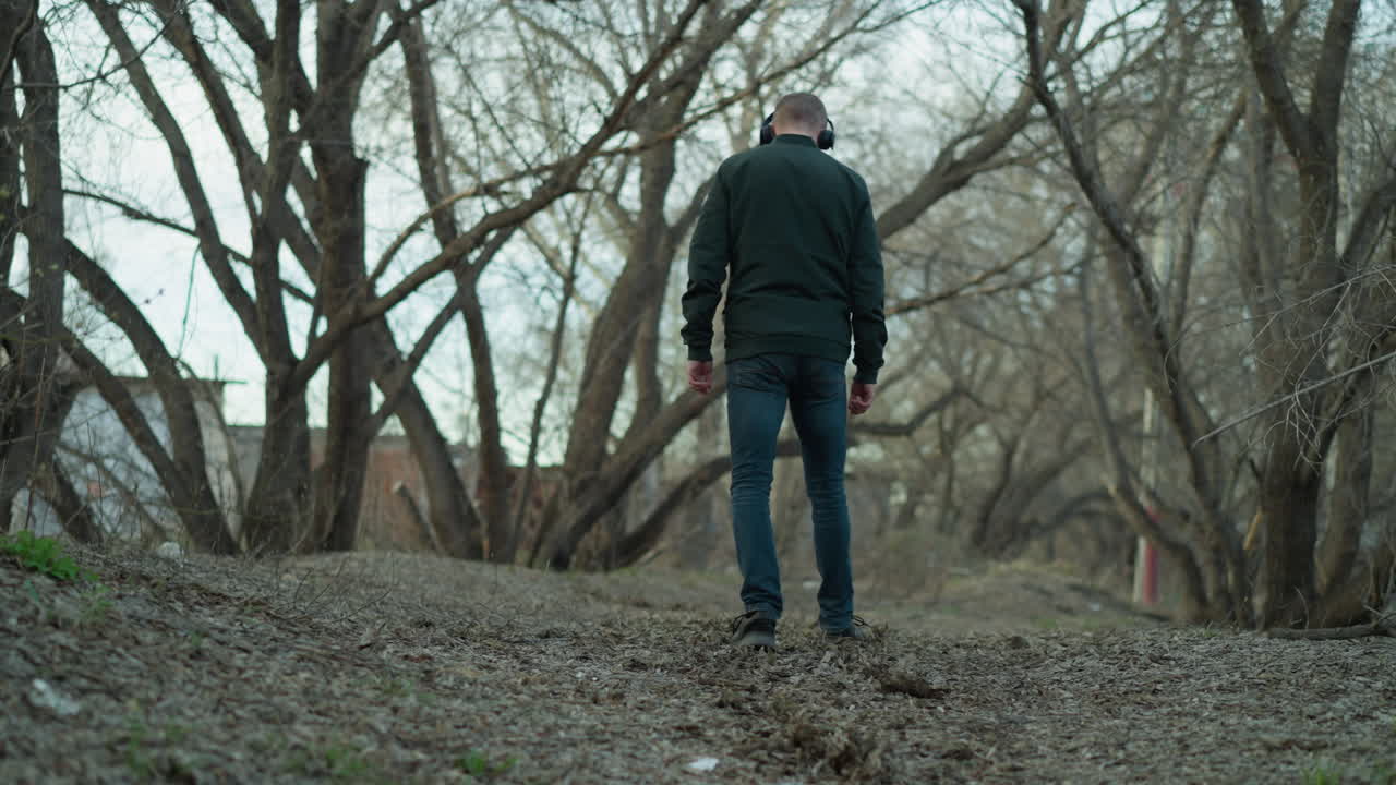 Man wearing a green jacket and headphones walking through a quiet forest, he is seen from behind, enjoying the peaceful surroundings of bare trees and a leaf-covered path