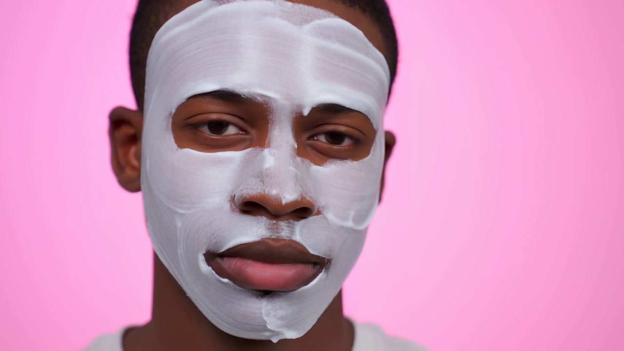Young Black Man Wearing White Mask, Closeup Studio Portrait Against Pink Background, Calm Expression