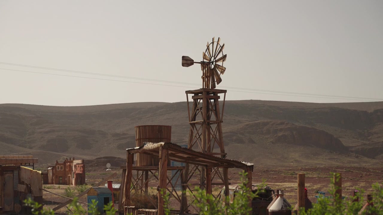 Rusty old windmill and wooden water tank structure rise above a scattered desert settlement and hills