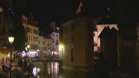 people walking along the thiou river at night with le palais de ille in annecy, france. - wide shot