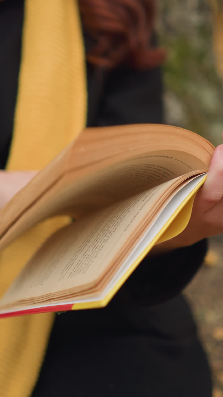 긴 스카프를 입고 책을 들고 페이지를 뒤집는 여성 손의 클로즈업, 가을 잎이 어져 자연에서 따뜻하고 평화로운 독서 분위기를 만니다.