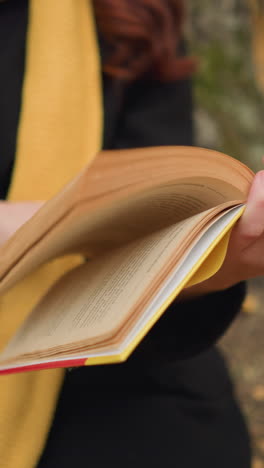 primer plano de la mano de la señora con una bufanda larga mientras sostiene el libro y hojea las páginas, hojas de otoño esparcidas por todas partes, creando una atmósfera de lectura cálida y pacífica en la naturaleza