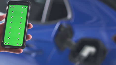 hombre cargando un coche eléctrico con un cable usando un teléfono de pantalla verde para monitorear el nivel de la batería