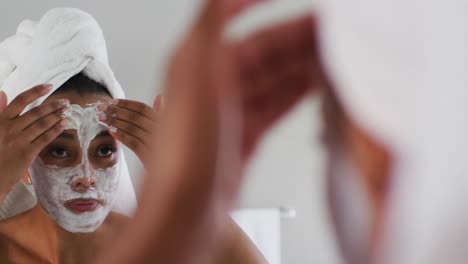 mujer afroamericana aplicando una máscara facial mirándose en el espejo en el baño