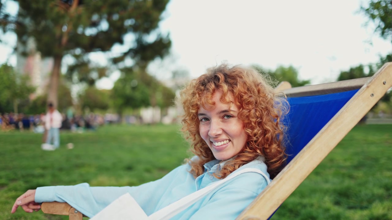 Giovane donna sorridente con capelli rossi ricci che si rilassa nel parco