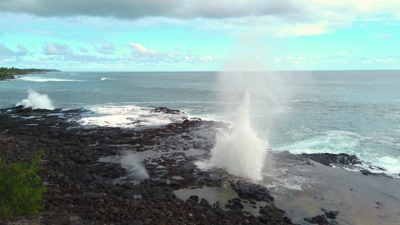 4K Hawaii Kauai static Spouting Horn blowhole going off in right center of frame as water swirls in hole in lava rock in left of frame with ocean in distance with partly cloudy sky