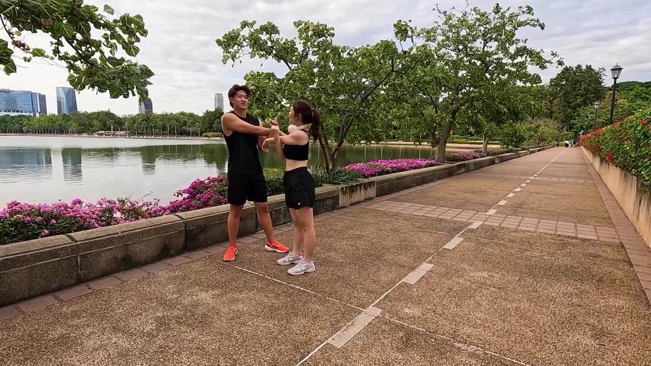 Pareja activa realiza estiramientos de brazos al aire libre junto a un lago con el horizonte de la ciudad de fondo durante el día