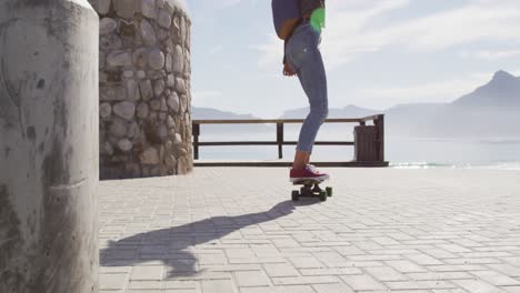 Rückansicht-Einer-Gemischtrassigen-Frau,-Die-Auf-Der-Sonnigen-Promenade-Am-Meer-Skateboard-Fährt