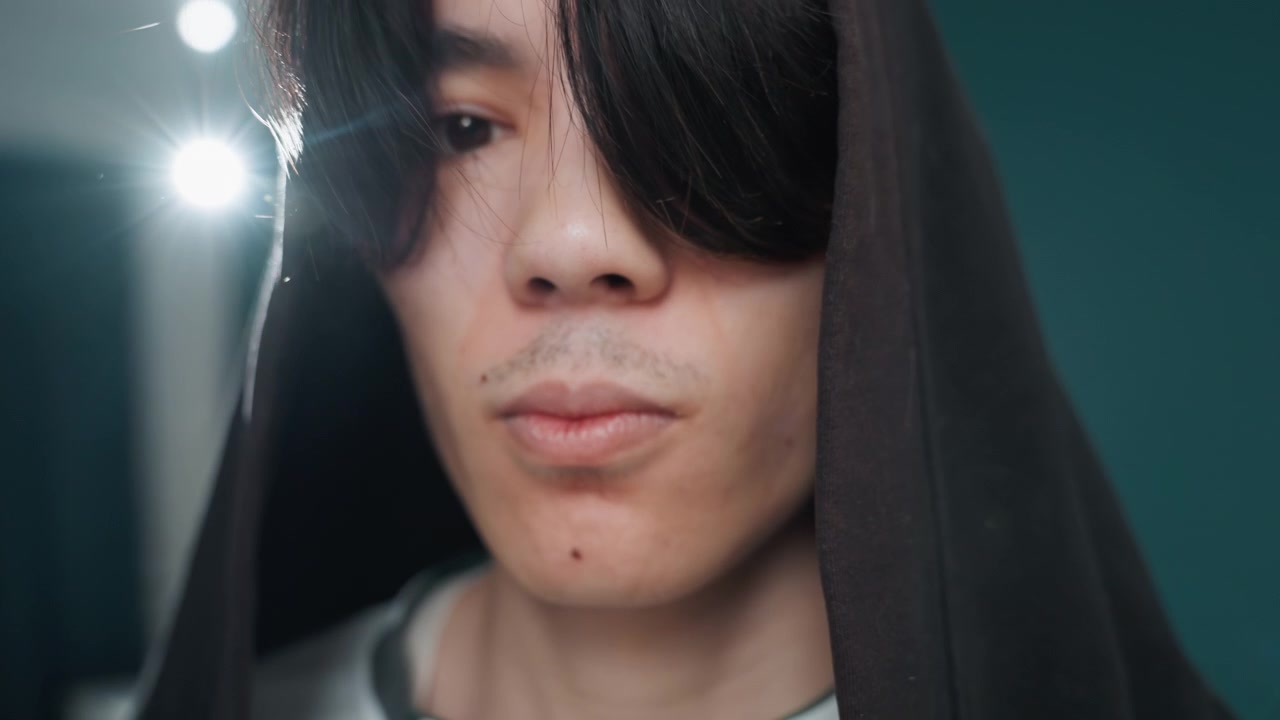 Potret berkerudung berhenti sejenak saat membersihkan rumah. Studi wajah jarak dekat dengan cahaya latar lembut, rambut jatuh menutupi mata, detail janggut tipis yang halus, ekspresi merenung, pencahayaan temaram, jeda tenang di rumah.