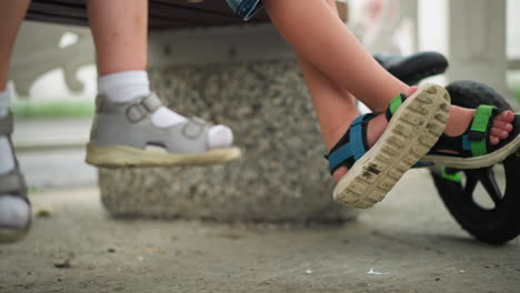 a close-up of two little children s legs, one crossing over the other, as they gently swing their feet back and forth while seated, a green bicycle is parked beside them