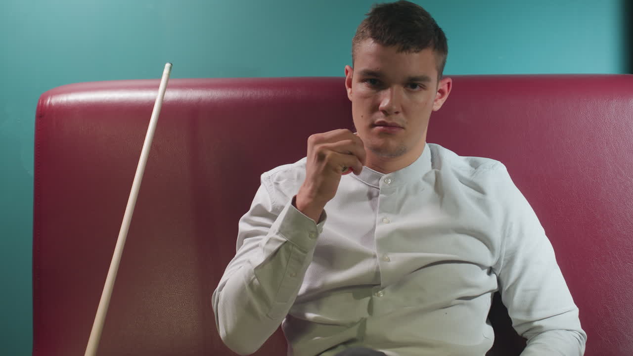 색 소파에 앉아있는 색 셔츠를 입은 남자의 클로즈업. 큐 스틱이 손으로 빌리어드 공을 던지면서 그의 에 기울어져 있습니다. 부드러운 조명은 그의 집중된 분위기를 강조합니다.