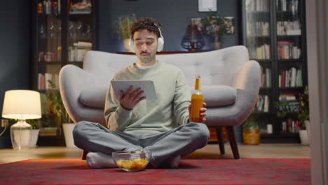 joven viendo una escena de película inesperada en una tableta mientras bebe refrescos y se sienta con las piernas cruzadas en el suelo en casa