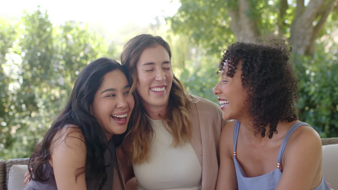 Amigas diversas sorrindo e trocando olhares, demonstrando afeto e se aproximando em um sofá de jardim.