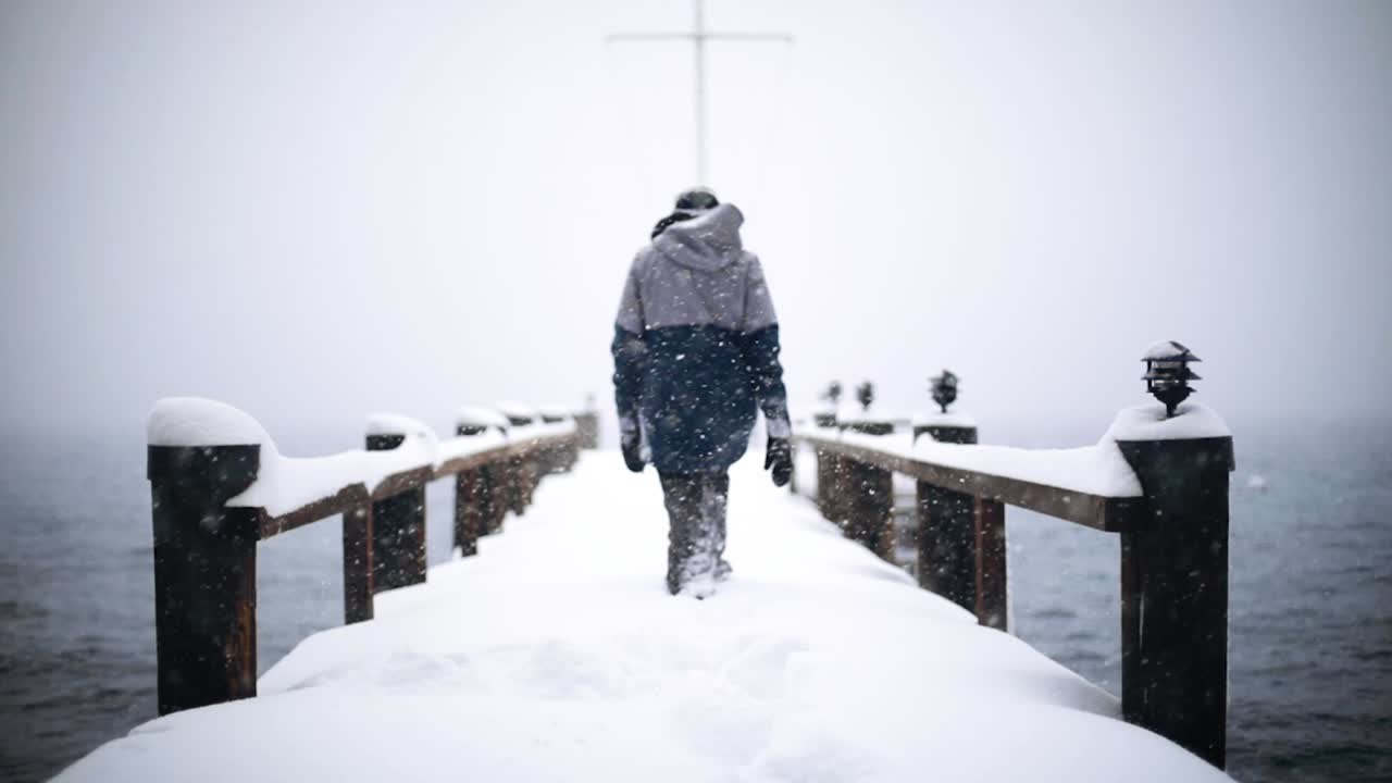 Person walking in slow motion through deep snow on a snow covered wooden pier over lake on a snowy, foggy day