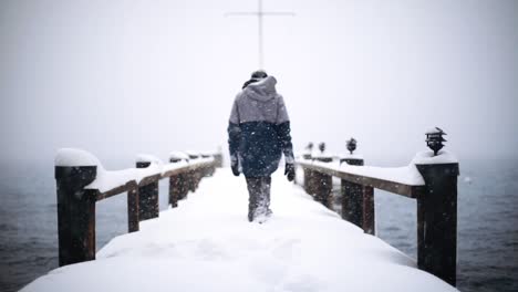 person walking in slow motion through deep snow on a snow covered wooden pier over lake on a snowy, foggy day