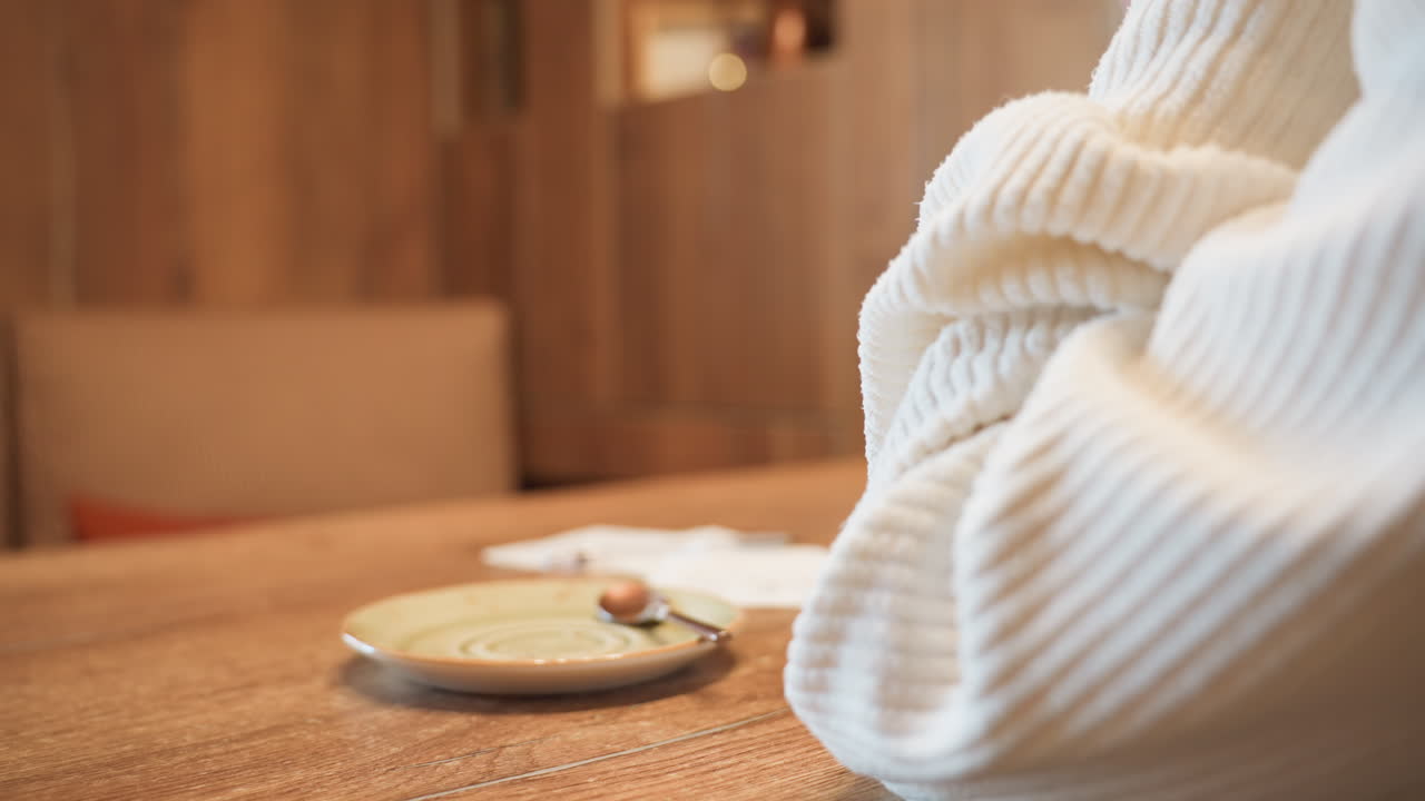 close up person in cozy ribbed white sweater lifting green ceramic cup from table beside small empty saucer and teaspoon in warm wood interior setting