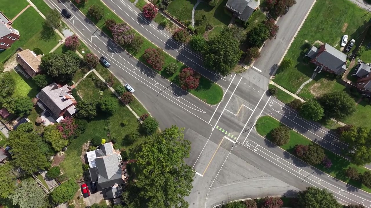 Quiet suburban street intersection with crosswalk markings winds past leafy Richmond, Virginia houses and colorful ornamental trees, single parked car visible from above. Aerial drone top down shot