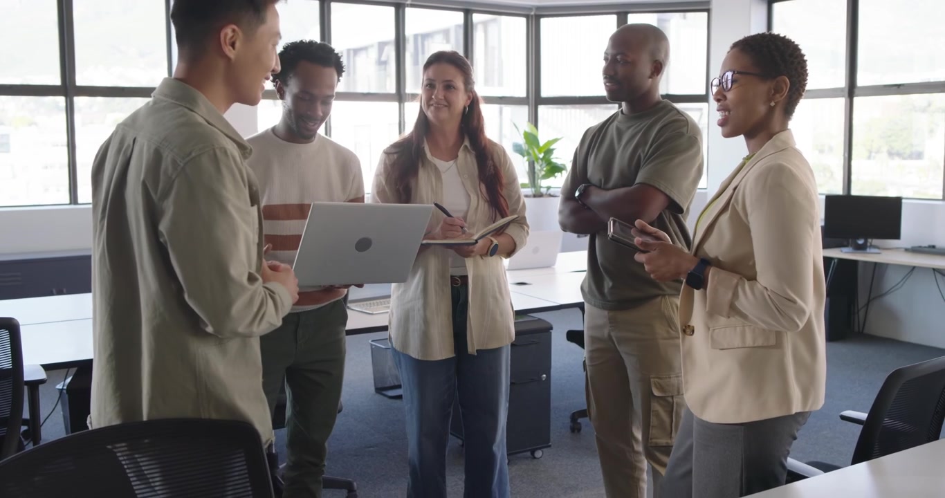 Colleghi diversi in un ufficio open space con raccoglitore e tablet, che mostrano un laptop, mentre due si uniscono per condividere appunti.