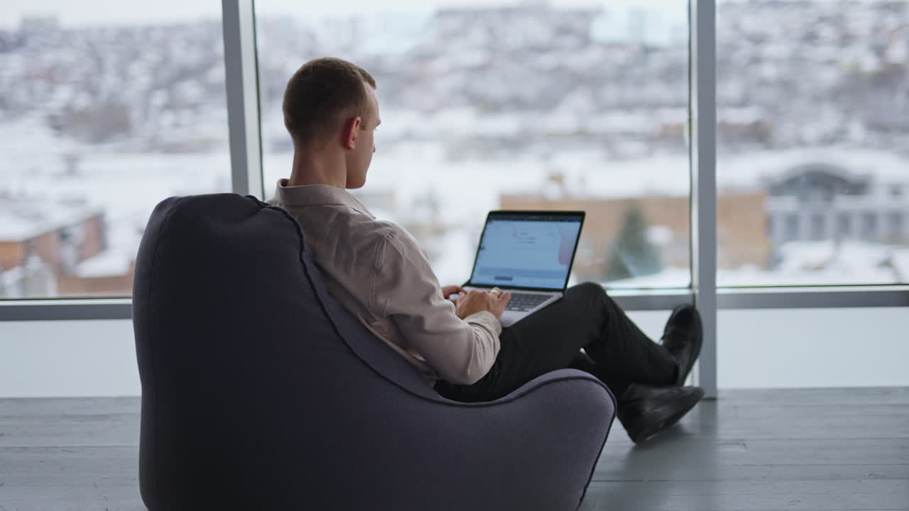 Busy male freelancer sits in bean bag chair with his back to camera. Man works on a laptop with graphs at the screen. Window with blurred cityscape at backdrop.