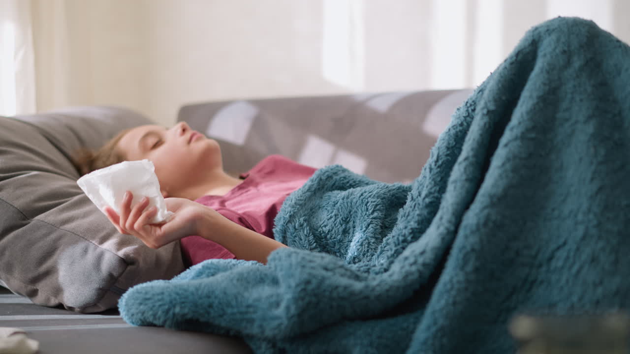 Student lying down on couch, sneezing into tissue. Covered with blanket, feeling unwell, tissue scattered around, representing flu symptoms