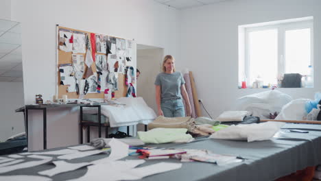 seamstress walks into workspace with apprentice, sets phone on cluttered table, picks design paper while apprentice watches attentively, creative fabrics, sewing tools, inspiration boards