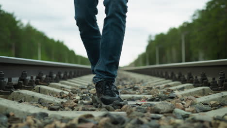 close leg view of someone wearing jeans and canvas shoes, walking tiredly on a railway with stones, the background includes a blur of trees and electric poles