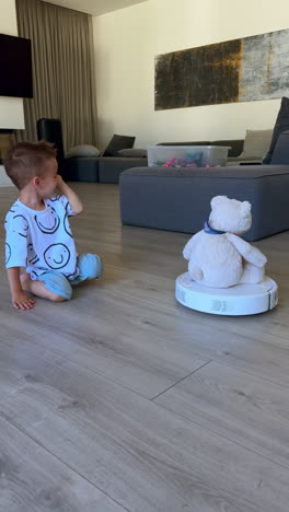 lovely caucasian baby watching the working robot cleaner. cute kid laughs looking at teddy bear riding the vacuum. vertical video.