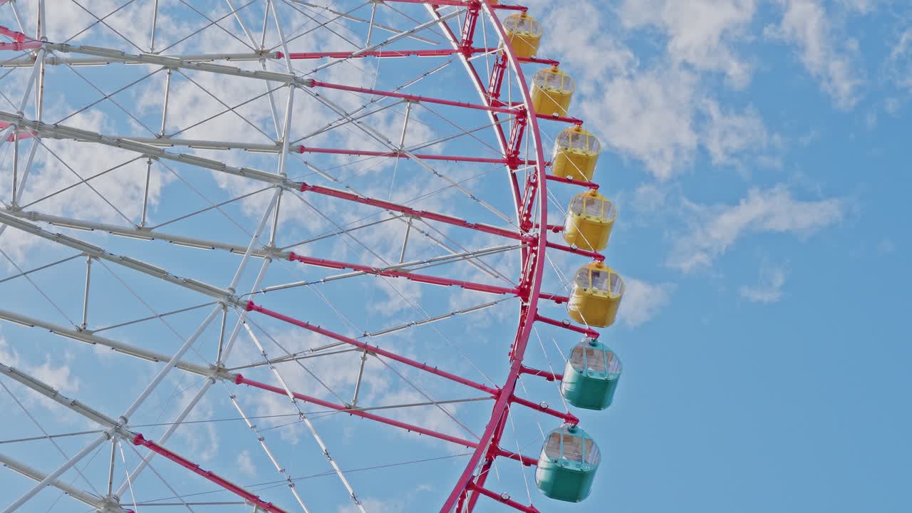 Close-up of yellow and green passenger cabins and the red spoke structure of a massive Ferris wheel against a cloudy blue sky