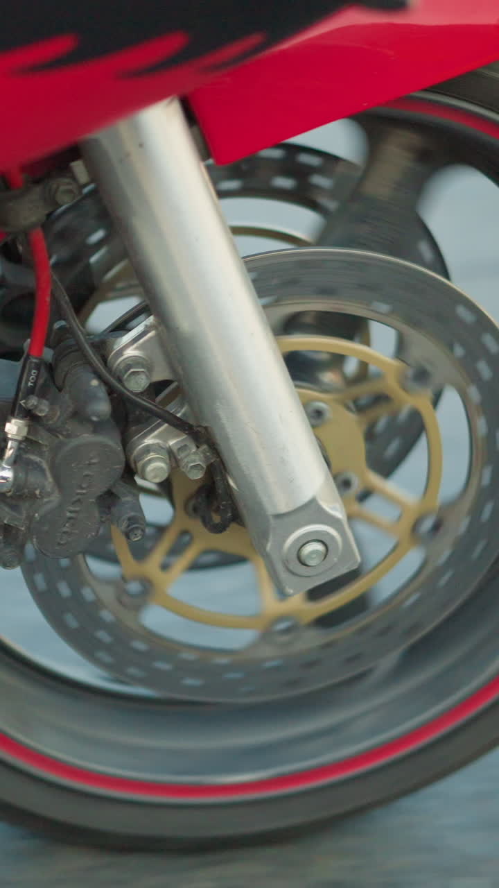 un primer plano de la rueda delantera de una motocicleta girando en un camino liso y asfalto, los componentes del neumático y los frenos están en foco