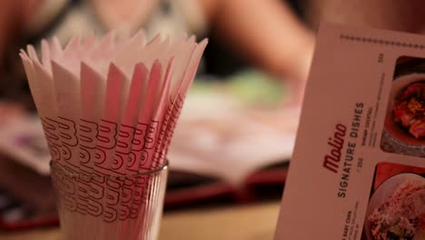 a person’s hands flip through a restaurant menu at a table, with a stack of pink paper napkins in focus. soft, warm lighting and shallow depth of field create an inviting atmosphere