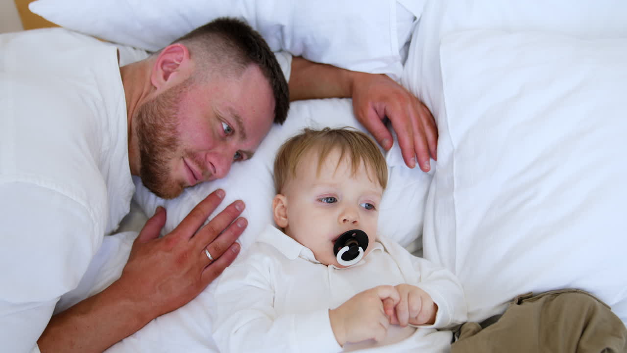 Loving dad lies near his baby looking at him with adoration. Toddler boy sucking a pacifier falling asleep. Top view.