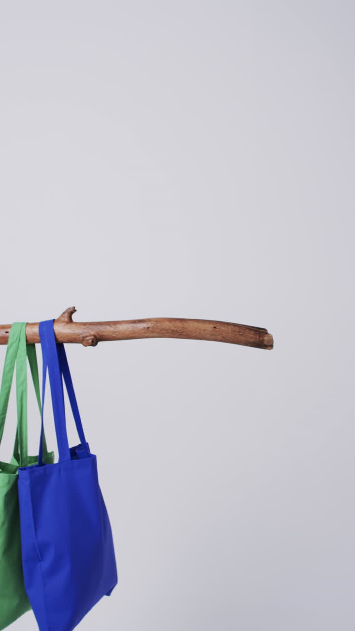 Vertical video of blue and green canvas bags hanging from branch with copy space on white background