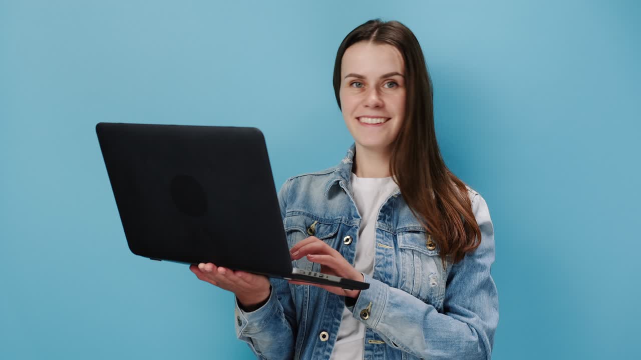 simpática joven caucásica de 20 años trabajando en una computadora portátil y mostrando el pulgar hacia arriba como un gesto, lleva una chaqueta de denim, aislada en la pared de fondo azul del estudio con espacio de copia para publicidad